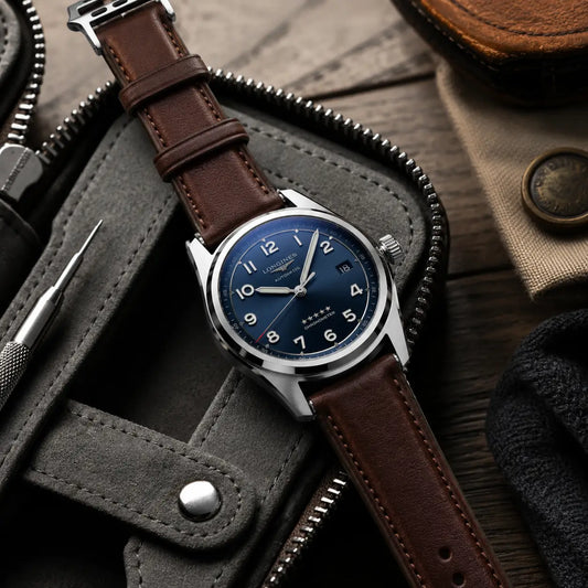 Luxury flat-lay product photograph of a blue-dial stainless steel watch paired with a smooth chocolate-brown leather strap and matching keeper loops. Styled on a charcoal travel case with leather accessories and a watch tool, the scene uses moody studio lighting for a sophisticated handcrafted aesthetic.