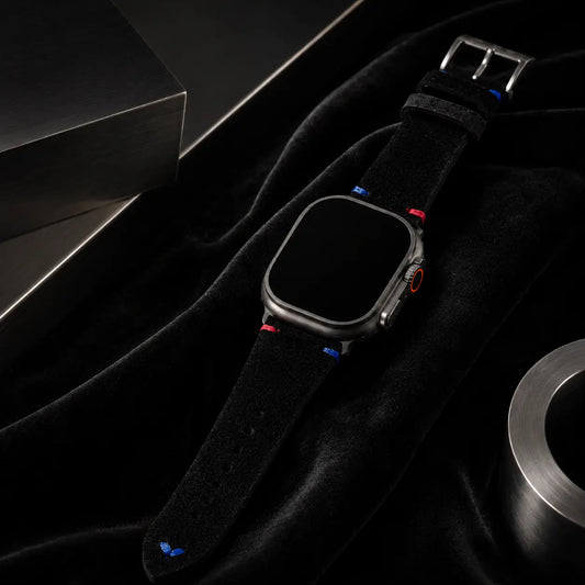 High-end luxury campaign image of an Apple Watch Ultra fitted with a black suede leather strap resting over rich black velvet and framed by sculptural brushed steel forms. Elegant light gradients reveal the soft material textures and colorful stitch details while maintaining a sophisticated monochrome aesthetic. The watch screen remains completely black.