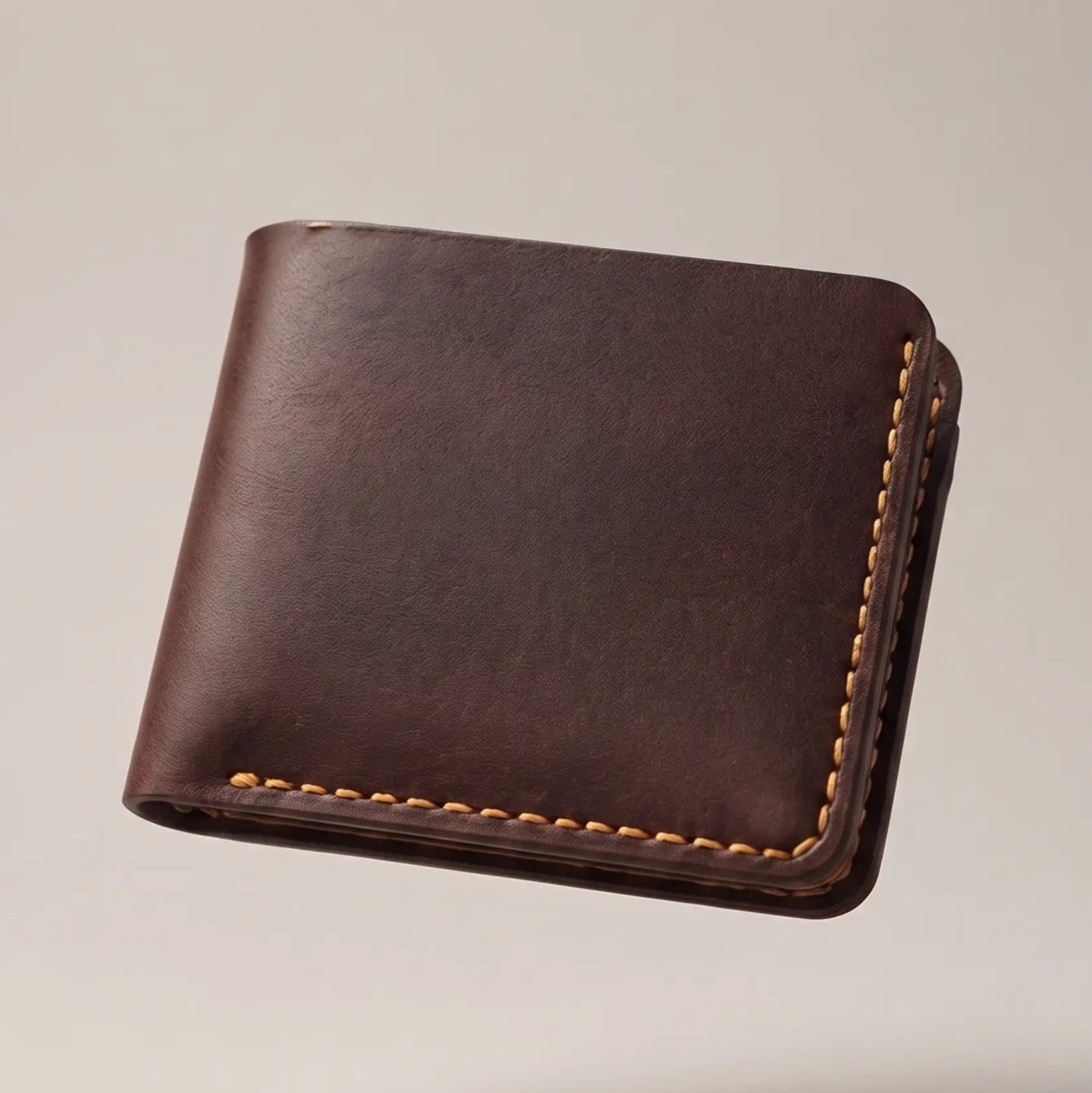 Dark brown full-grain leather bifold wallet with tan contrast stitching floating against a clean beige background, highlighting its slim profile, minimalist design, and handcrafted finish.