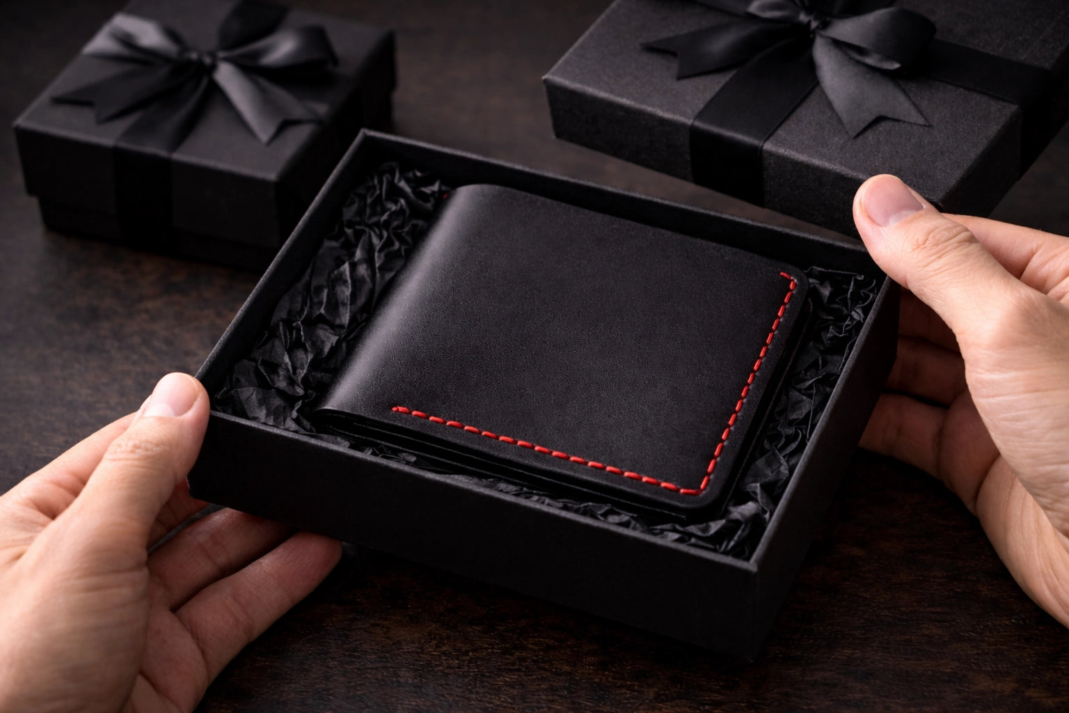 Black leather wallet with red stitching held by hands, displayed in a black box on a dark surface.