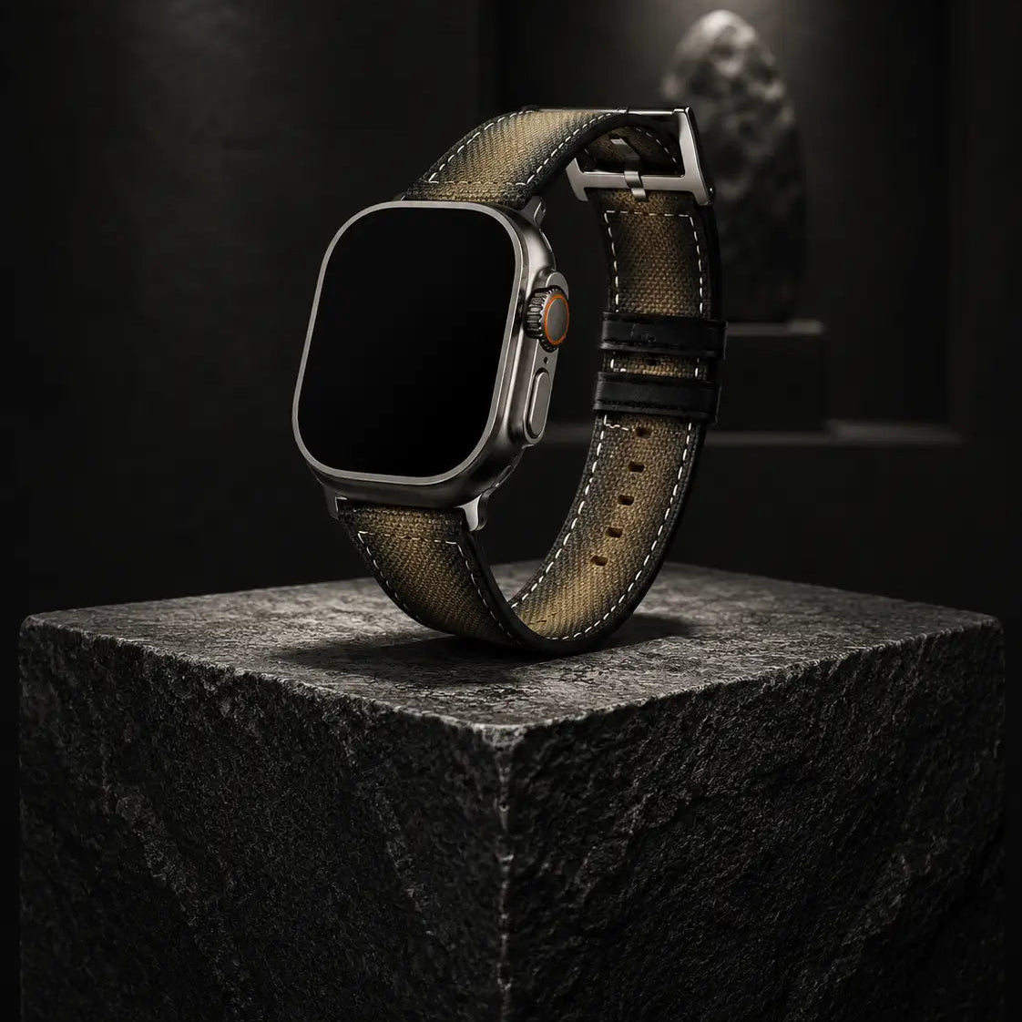 Luxury editorial product photograph of an Apple Watch Ultra fitted with a khaki canvas strap, standing upright on a sculptural black stone pedestal inside a dark gallery-inspired setting. Museum-style spotlighting creates dramatic contrast and highlights the woven strap texture, precision stitching, and titanium case finish. The watch screen is completely off and pure black, reinforcing a refined quiet-luxury aesthetic.