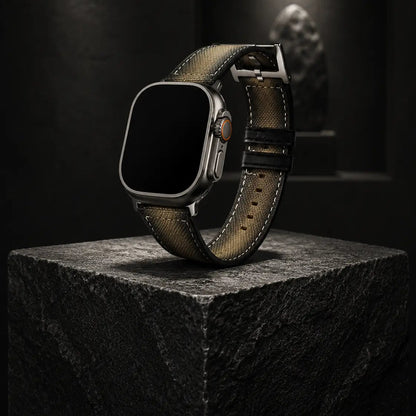 Luxury editorial product photograph of an Apple Watch Ultra fitted with a khaki canvas strap, standing upright on a sculptural black stone pedestal inside a dark gallery-inspired setting. Museum-style spotlighting creates dramatic contrast and highlights the woven strap texture, precision stitching, and titanium case finish. The watch screen is completely off and pure black, reinforcing a refined quiet-luxury aesthetic.