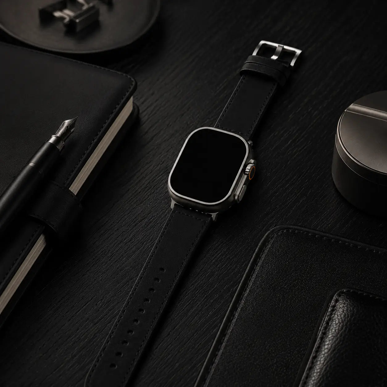 Apple Watch Ultra fitted with a black leather strap resting on a matte black oak desk beside a fountain pen, leather notebook, and refined desk accessories. Soft directional lighting highlights the smooth leather texture and brushed titanium case, creating an understated luxury editorial aesthetic with the screen completely black.