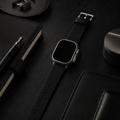 Apple Watch Ultra fitted with a black leather strap resting on a matte black oak desk beside a fountain pen, leather notebook, and refined desk accessories. Soft directional lighting highlights the smooth leather texture and brushed titanium case, creating an understated luxury editorial aesthetic with the screen completely black.