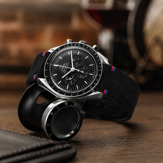 Luxury chronograph watch with a black suede-style strap featuring red and blue accent stitching, displayed on a dark wooden desk beside a black leather notebook, magnifying loupe, and warm ambient background for a premium editorial look.