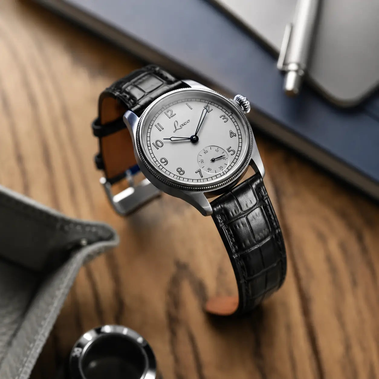 Premium black alligator grain leather watch strap with large square scales, paired with a Laco pilot watch on a grey notebook and wooden desk.