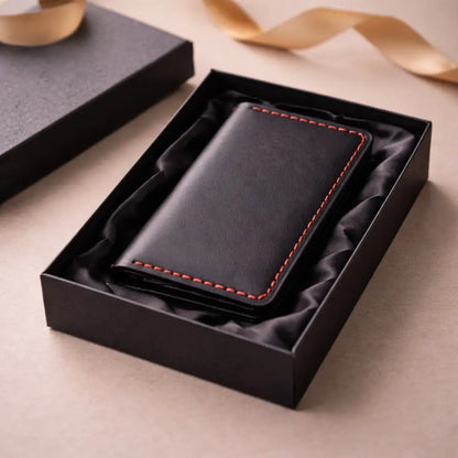 Black leather wallet with red stitching neatly presented inside an open black gift box lined with soft fabric, styled as a premium gifting setup with a ribbon in the background.