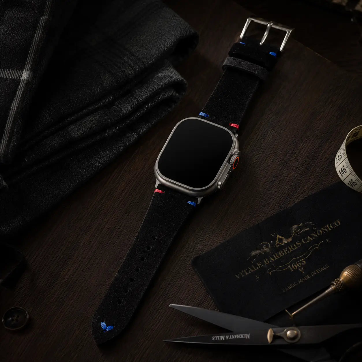 Luxury editorial product photograph of an Apple Watch Ultra fitted with a black suede leather strap featuring subtle blue and red stitch accents. The watch is arranged diagonally on a dark walnut tailoring table beside folded black cashmere, tailoring scissors, and atelier accessories. Soft cinematic lighting highlights the texture of the strap while the watch display remains completely black.