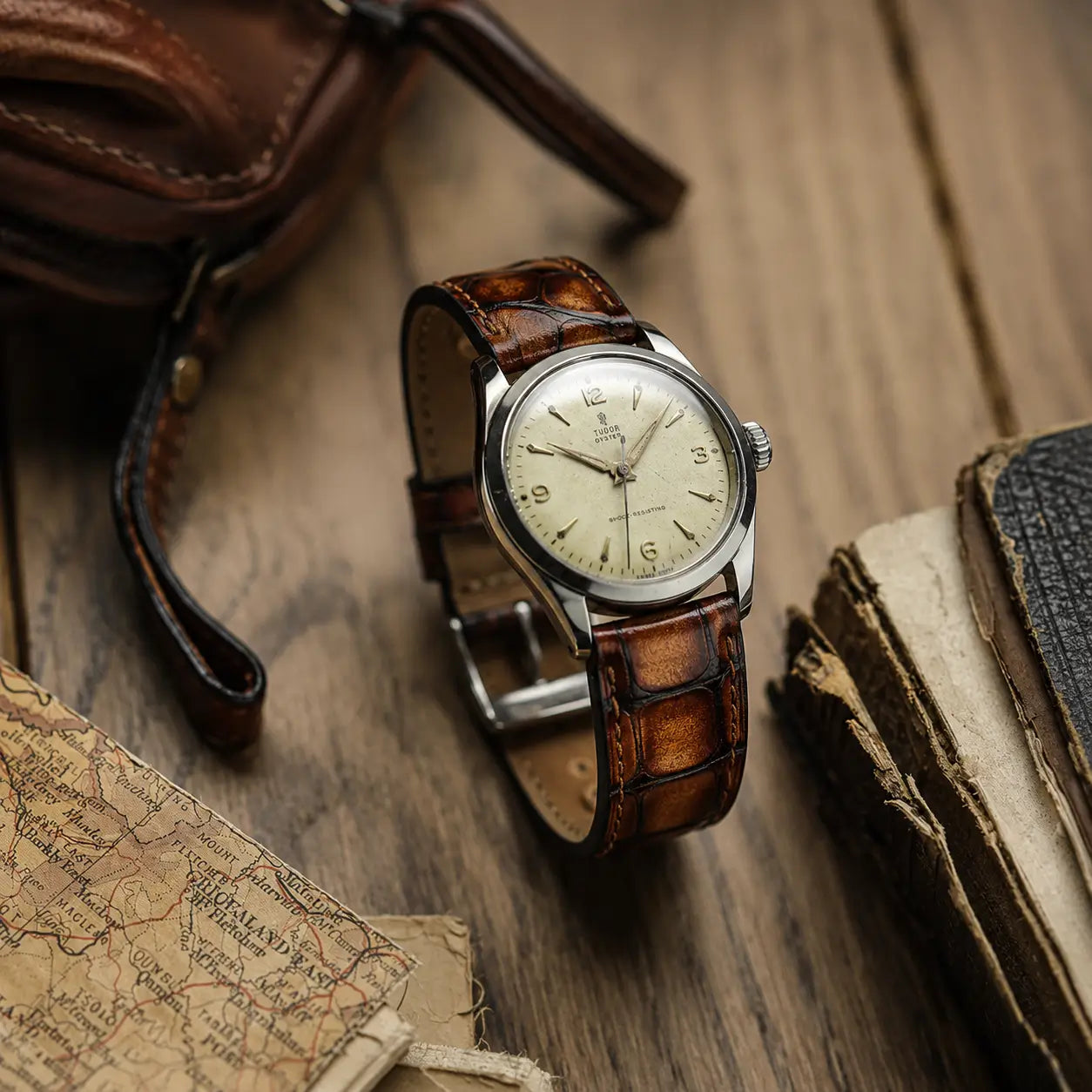 Honey brown crocodile grain leather watch strap with hand-stitched edges, paired with a vintage Tudor Oyster watch on a rustic wooden table with old maps.