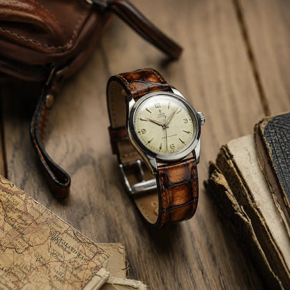 Honey brown crocodile grain leather watch strap with hand-stitched edges, paired with a vintage Tudor Oyster watch on a rustic wooden table with old maps.