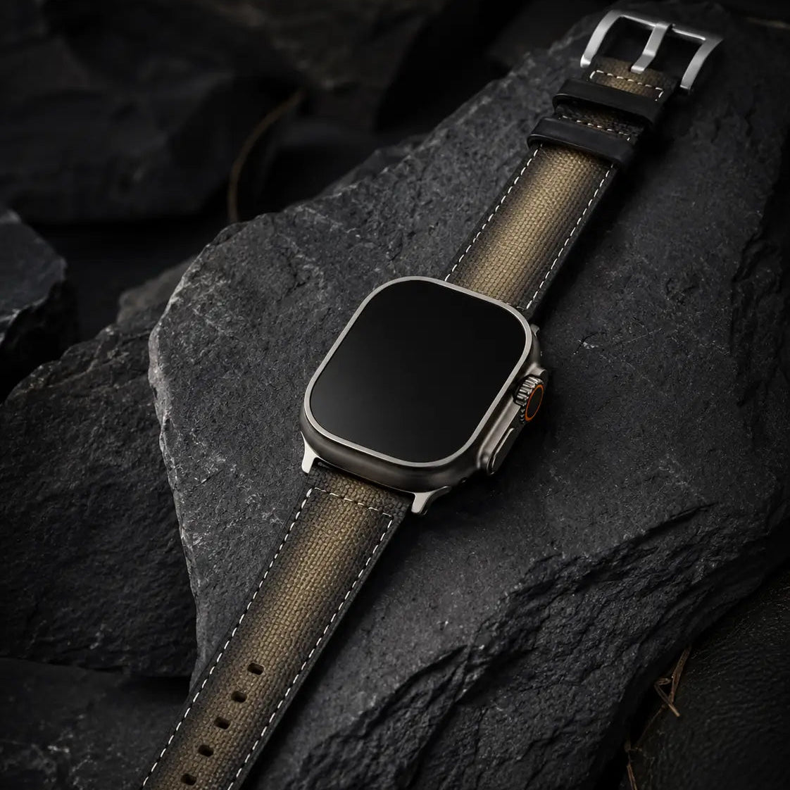 High-end luxury watch-style product photograph showing an Apple Watch Ultra laid across textured volcanic stone with the khaki canvas strap fully extended. Rich, controlled lighting emphasizes material craftsmanship, subtle fabric weave, and contrast stitching while maintaining the strap’s original khaki color tone. The watch face is completely off with a seamless black glass finish against a dark, premium environment.