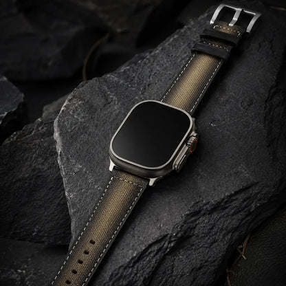 High-end luxury watch-style product photograph showing an Apple Watch Ultra laid across textured volcanic stone with the khaki canvas strap fully extended. Rich, controlled lighting emphasizes material craftsmanship, subtle fabric weave, and contrast stitching while maintaining the strap’s original khaki color tone. The watch face is completely off with a seamless black glass finish against a dark, premium environment.