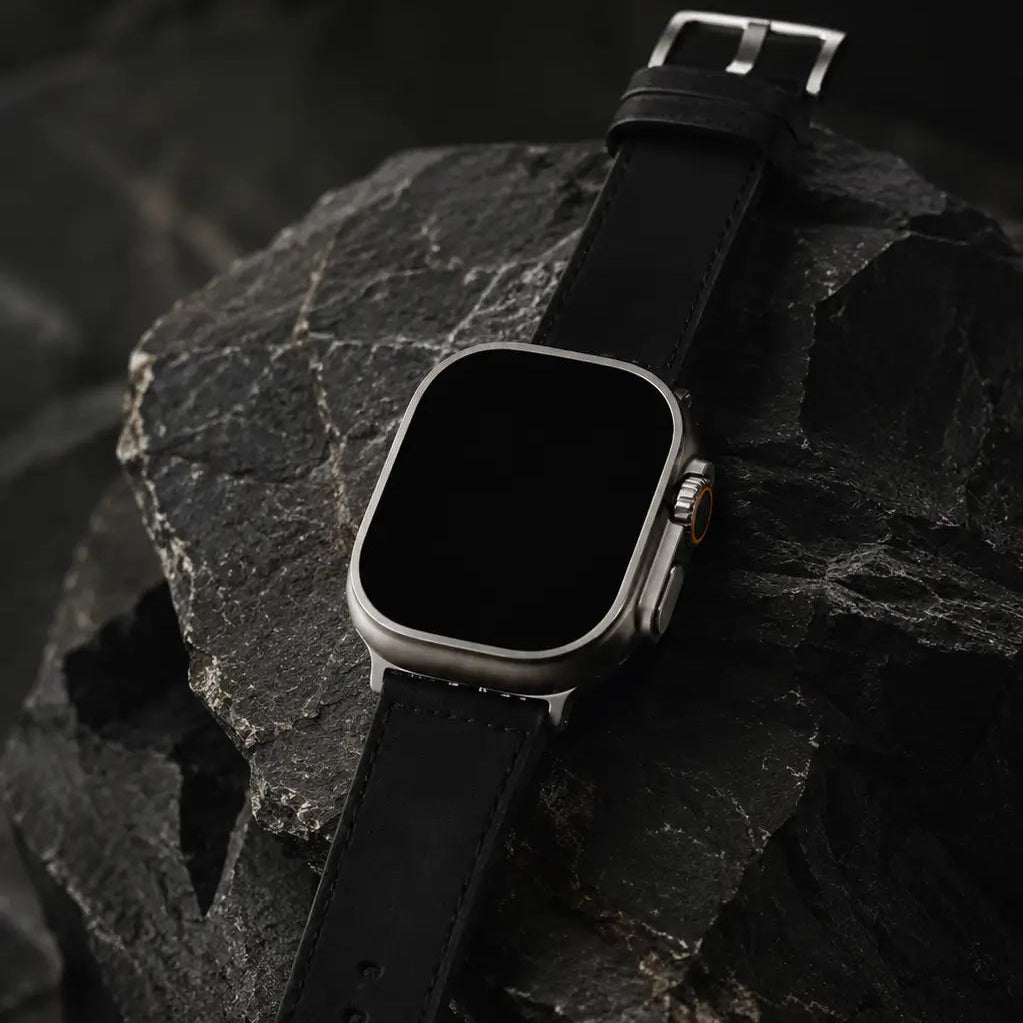 Apple Watch Ultra displayed on sculptural dark stone with the black leather strap extending across layered rock surfaces. Dramatic cinematic lighting emphasizes the premium materials, subtle stitching, and matte titanium finish. The screen remains fully off for a minimalist luxury campaign look.