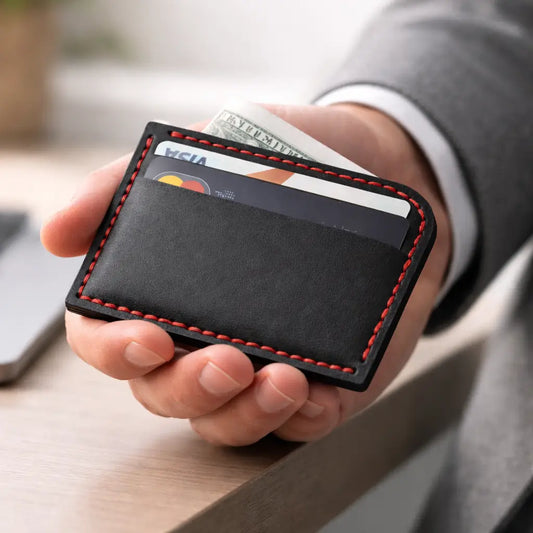 Hand holding a black leather cardholder with red stitching, containing credit cards and cash, over a desk surface.