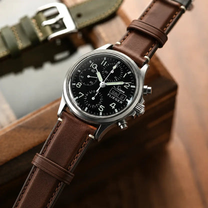 Close-up lifestyle shot of a stainless steel pilot chronograph watch with a rich chocolate-brown leather strap featuring tonal stitching and Apple Watch–style adapters. The watch rests diagonally on a warm wooden surface with a softly blurred olive strap in the background, creating a refined, premium editorial look.