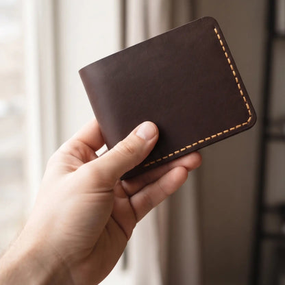 Hand holding a handcrafted dark brown leather bifold wallet with tan contrast stitching near a window, emphasizing the wallet's premium leather texture, slim silhouette, and everyday carry style.