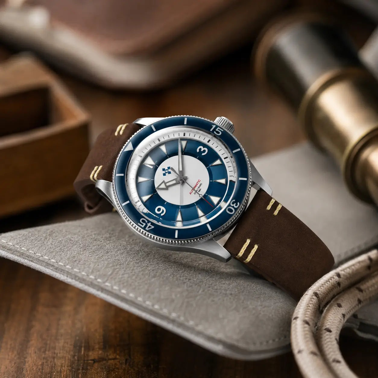 Luxury dive-style wristwatch with a deep blue bezel and silver case displayed on a grey leather surface. The watch is fitted with a dark brown leather strap featuring contrast hand stitching, surrounded by warm-toned props including a brass telescope, wooden tray, and coiled rope for an editorial lifestyle setting.