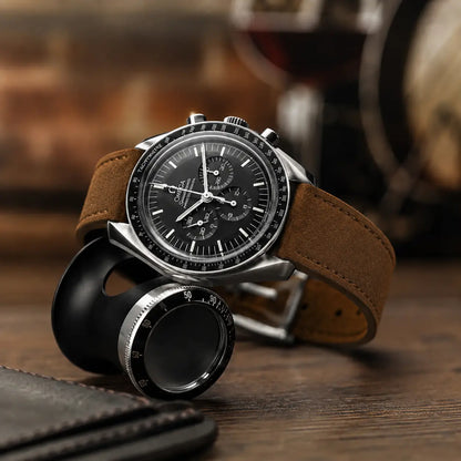 Vintage-inspired luxury watch with a warm brown suede leather strap held in hand, styled with leather accessories and watchmaking tools on a dark wooden surface for an editorial lifestyle look.