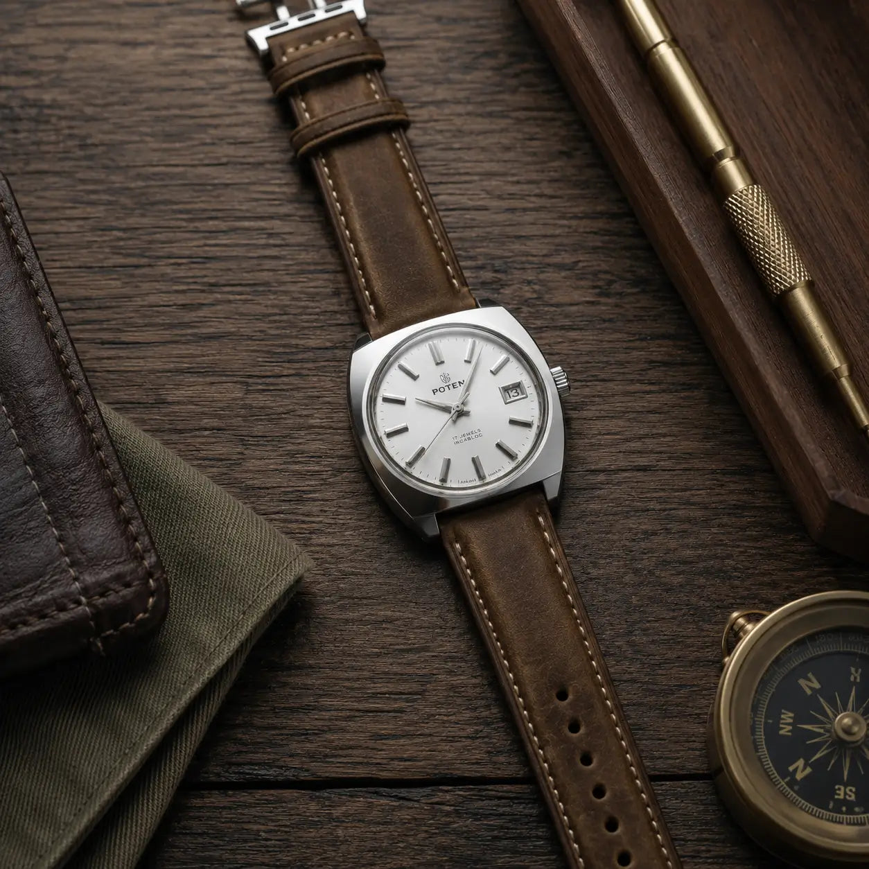 Premium lifestyle watch photograph featuring a vintage-inspired stainless steel wristwatch with a distressed brown leather strap and contrast stitching, laid flat on a dark wooden surface. The watch has a clean white dial with silver hour markers and a date window. Surrounding props include a textured leather wallet, folded olive fabric, a brass precision tool in a wooden tray, and a brass compass, creating a refined editorial aesthetic with warm lighting and shallow depth of field.
