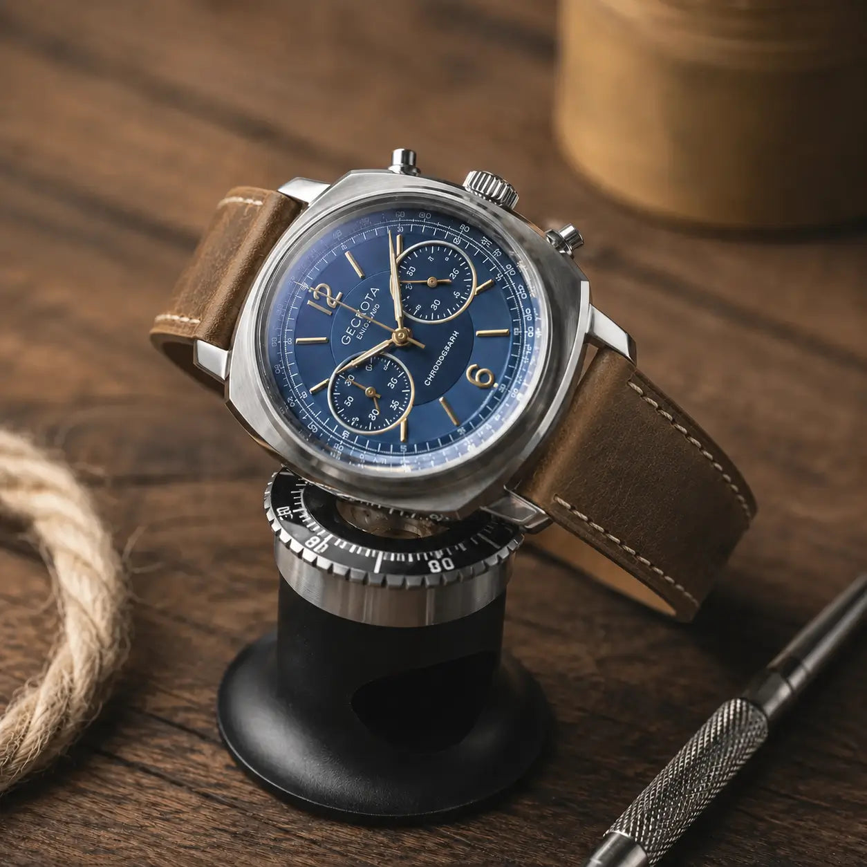 Editorial product photograph of a stainless steel chronograph watch with a deep blue dial and gold-tone markers, displayed on a black watch stand atop a rustic wooden surface. The watch is fitted with a distressed brown leather strap featuring contrast stitching and a vintage finish. Soft warm lighting highlights the brushed metal case and textured leather, while blurred background elements including rope, a metallic container, and a precision tool create a refined workshop-inspired luxury aesthetic.