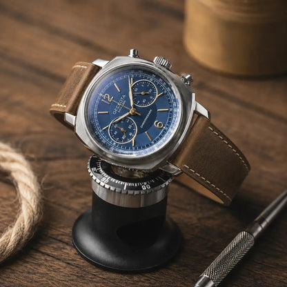 Editorial product photograph of a stainless steel chronograph watch with a deep blue dial and gold-tone markers, displayed on a black watch stand atop a rustic wooden surface. The watch is fitted with a distressed brown leather strap featuring contrast stitching and a vintage finish. Soft warm lighting highlights the brushed metal case and textured leather, while blurred background elements including rope, a metallic container, and a precision tool create a refined workshop-inspired luxury aesthetic.