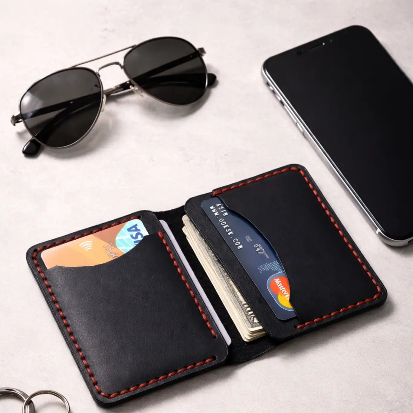 A handcrafted black leather bifold card holder with vibrant red contrast stitching, shown open to reveal credit cards and folded cash, arranged on a light grey surface with everyday essentials including aviator sunglasses, a smartphone, and a car key fob.