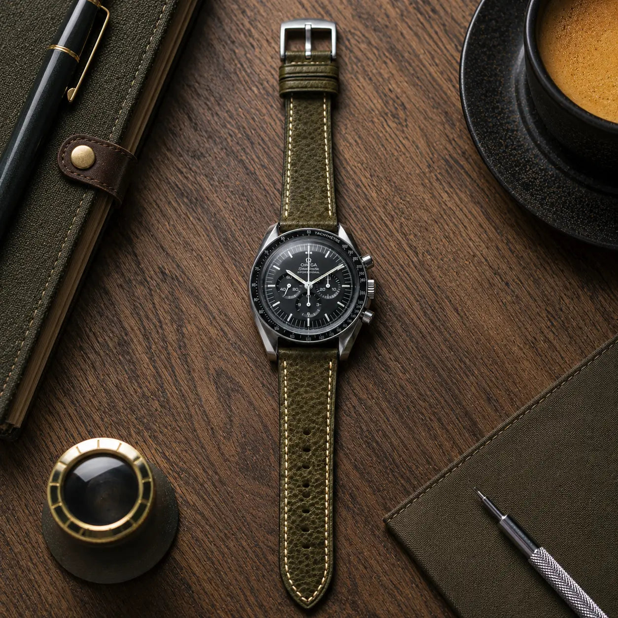 20mm Olive green leather watch strap fitted on Omega Speedmaster, premium handcrafted watch band with textured grain and classic stitching on wooden desk.