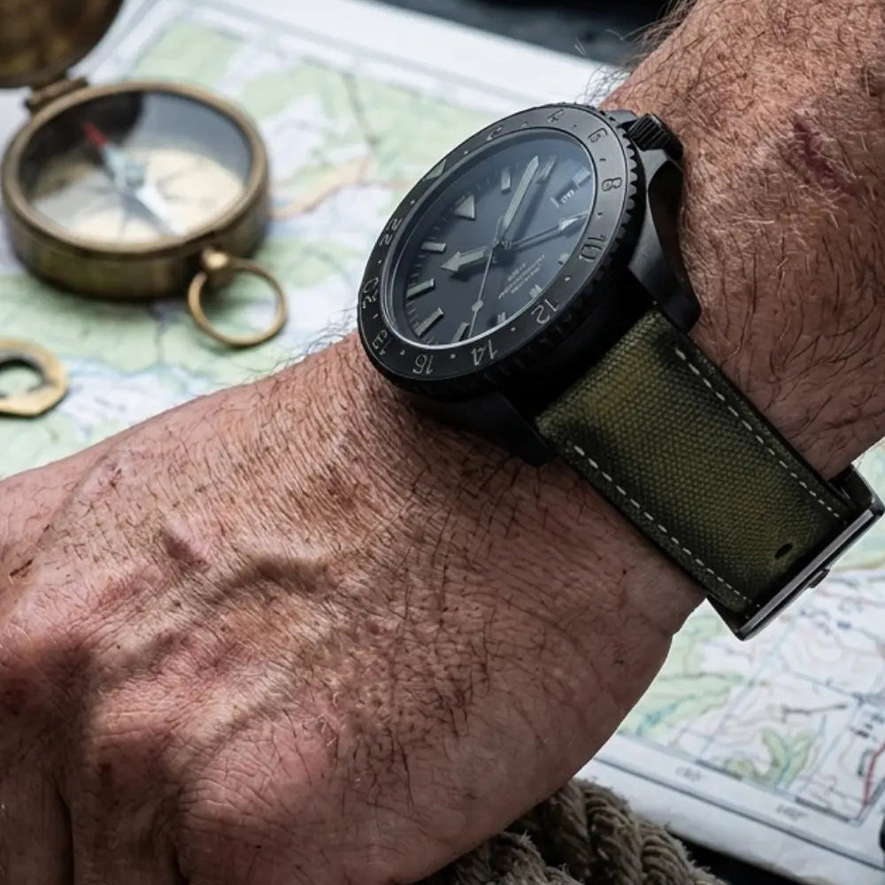 Black GMT wristwatch with a khaki canvas watch strap worn on the wrist above a vintage map, rope, and brass compass, highlighting a durable military-inspired design for outdoor adventures.