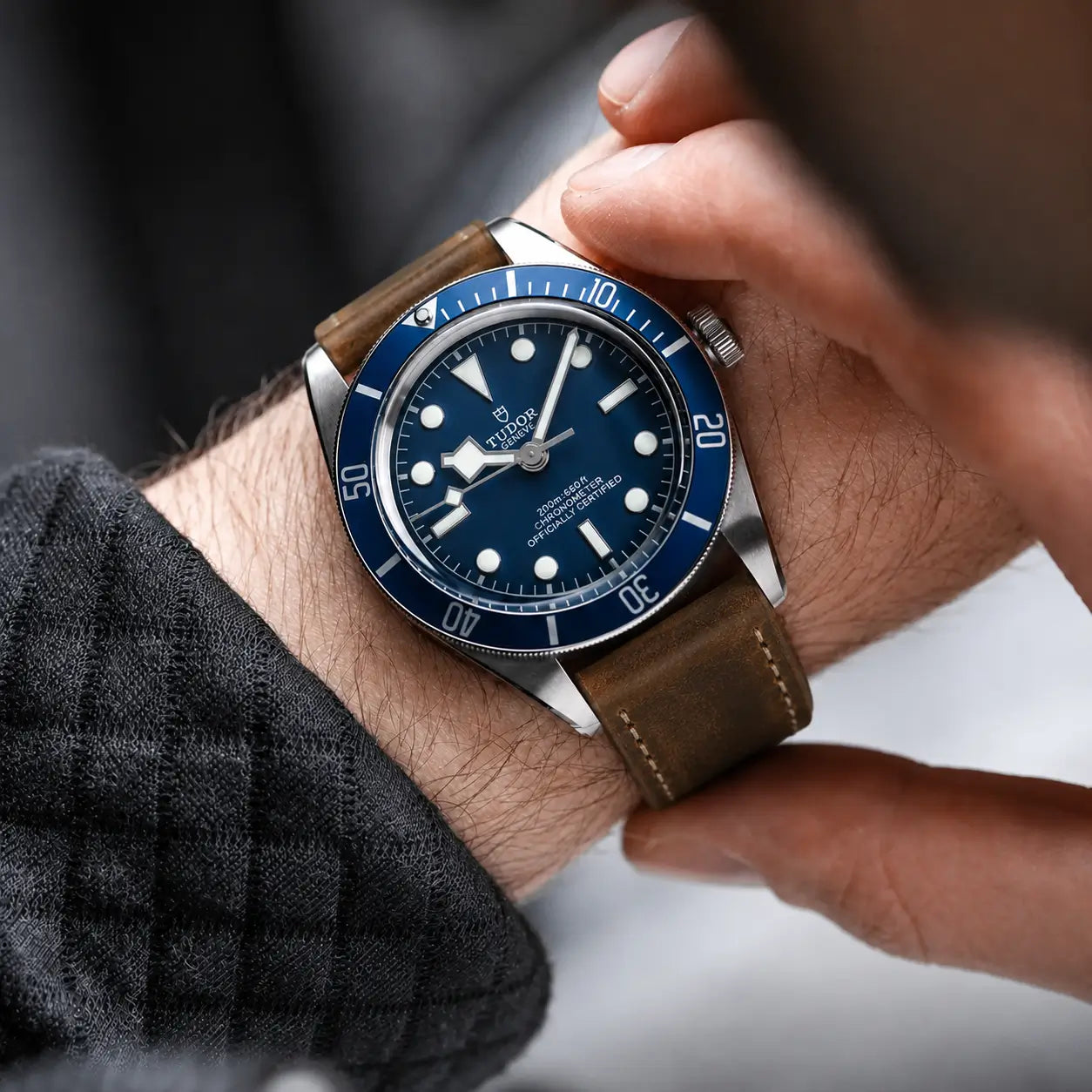 Luxury lifestyle close-up of a stainless steel diver’s watch with a deep blue dial and bezel, worn on the wrist with a distressed brown leather strap featuring contrast stitching. A hand adjusts the watch while soft natural lighting highlights the brushed metal case, rich leather texture, and fine details. The background is softly blurred, creating a premium editorial and modern watch photography aesthetic.