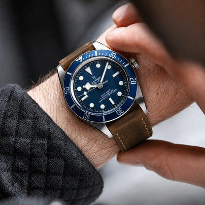 Luxury lifestyle close-up of a stainless steel diver’s watch with a deep blue dial and bezel, worn on the wrist with a distressed brown leather strap featuring contrast stitching. A hand adjusts the watch while soft natural lighting highlights the brushed metal case, rich leather texture, and fine details. The background is softly blurred, creating a premium editorial and modern watch photography aesthetic.