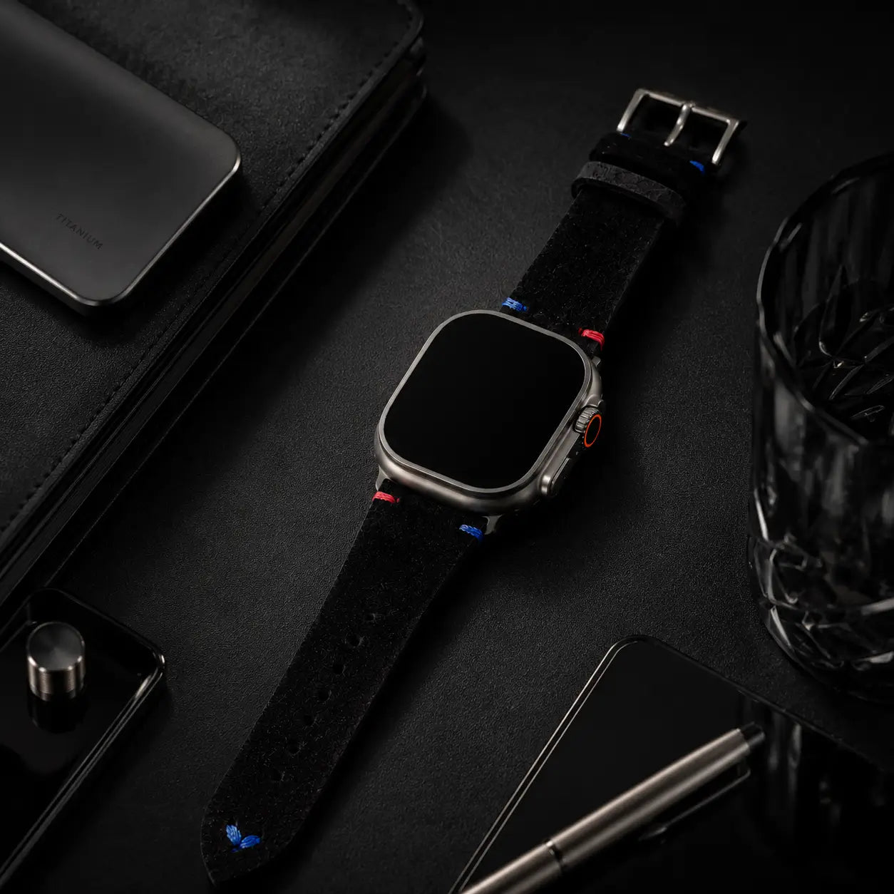 Premium luxury still-life of an Apple Watch Ultra with a black suede strap displayed across a matte black executive desk. Surrounded by smoked glass, titanium accessories, and refined office elements, the composition uses low-key lighting and deep shadows to emphasize modern executive luxury. The screen is fully off and pure black.