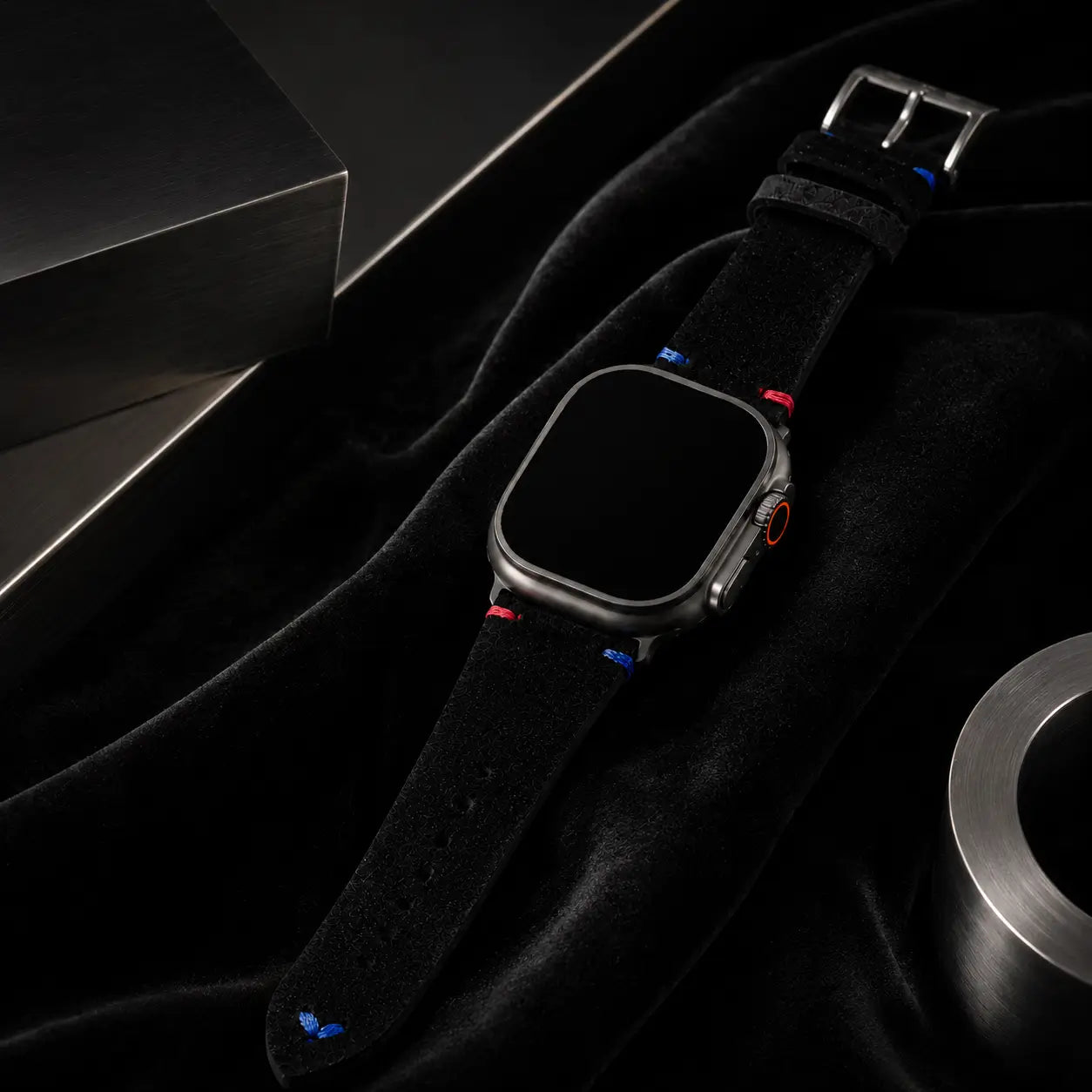High-end luxury campaign image of an Apple Watch Ultra fitted with a black suede leather strap resting over rich black velvet and framed by sculptural brushed steel forms. Elegant light gradients reveal the soft material textures and colorful stitch details while maintaining a sophisticated monochrome aesthetic. The watch screen remains completely black.