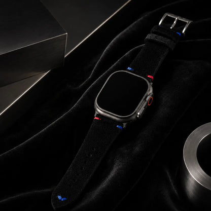 High-end luxury campaign image of an Apple Watch Ultra fitted with a black suede leather strap resting over rich black velvet and framed by sculptural brushed steel forms. Elegant light gradients reveal the soft material textures and colorful stitch details while maintaining a sophisticated monochrome aesthetic. The watch screen remains completely black.