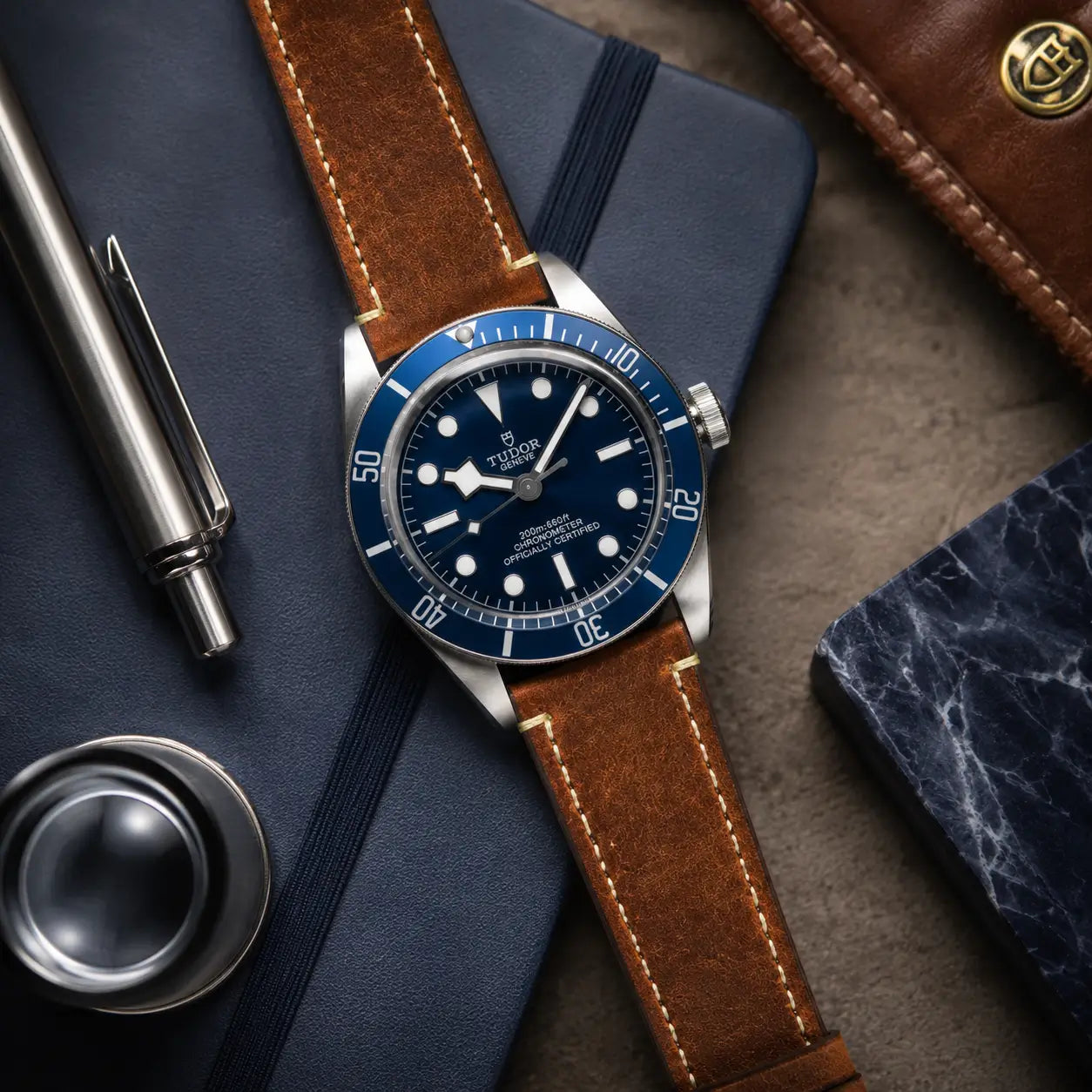 Luxury flat lay image of a blue diver-style watch featuring a natural tan crazy horse leather strap, styled on a navy notebook with metallic accessories and rich textures in a high-end studio setting.