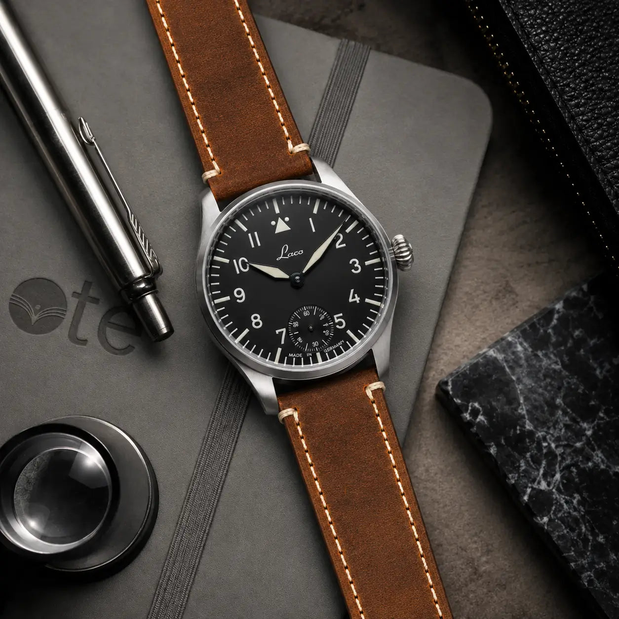 Premium flat lay product photograph of a black dial pilot watch paired with a natural tan leather watch strap, arranged on a charcoal notebook with a metal pen, magnifier, and marble accent for a refined editorial look.