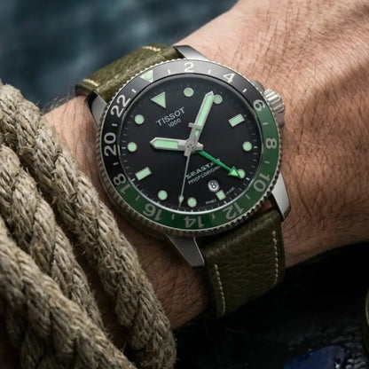Close-up of a Tissot GMT watch worn with an olive green full-grain leather watch strap on the wrist, paired with a knitted sweater for a rugged outdoor lifestyle and everyday wear.
