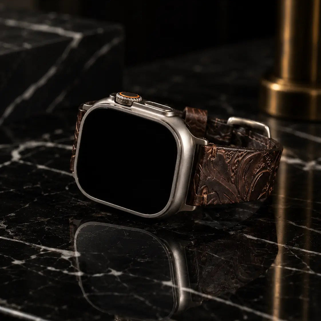 Luxury still-life of Apple Watch Ultra wearing an embossed dark brown leather strap positioned on polished black marble with a soft mirrored reflection. Dramatic studio lighting and bronze accents emphasize premium materials and refined watch styling with the screen turned off.