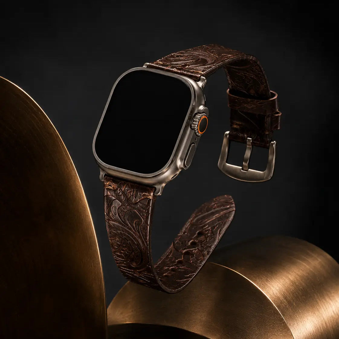 Apple Watch Ultra with embossed dark brown leather strap suspended above sculptural brushed bronze forms in a dark editorial studio. Subtle reflections and directional edge lighting showcase the detailed leather craftsmanship and metallic buckle against a deep charcoal background with the screen fully black.