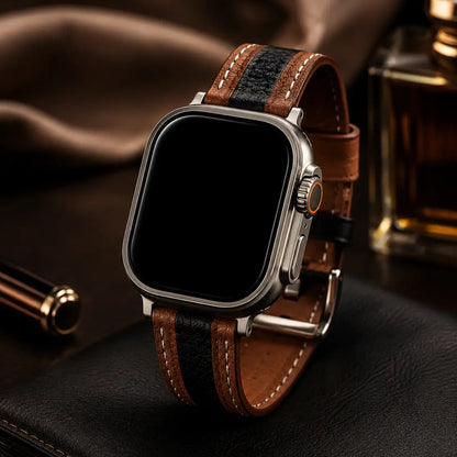 Apple Watch Ultra fitted with a two-tone brown and black leather strap standing upright on a black leather surface. Warm cinematic lighting highlights the textured leather, white contrast stitching, and brushed titanium case. Soft-focus luxury accessories and amber-toned glass elements in the background create an elegant premium editorial aesthetic. The watch screen is completely off and black.