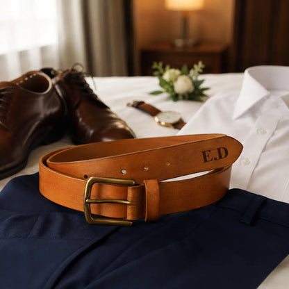 Cognac brown leather men’s belt with engraved initials E.D. styled with formal trousers and leather shoes on a bed.