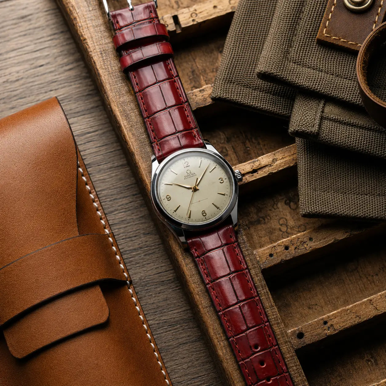 Deep cherry red alligator grain leather watch band with color-matched stitching, pictured on a silver-dial Baltic watch inside a grey suede travel pouch.