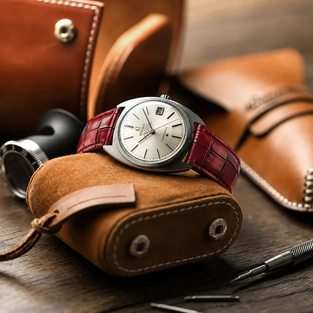 Ruby red crocodile embossed leather watch strap paired with a Baltic watch, displayed on a wooden table alongside a fountain pen, a watchmaker's loupe.