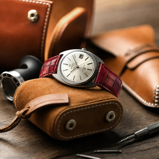 Ruby red crocodile embossed leather watch strap paired with a Baltic watch, displayed on a wooden table alongside a fountain pen, a watchmaker's loupe.