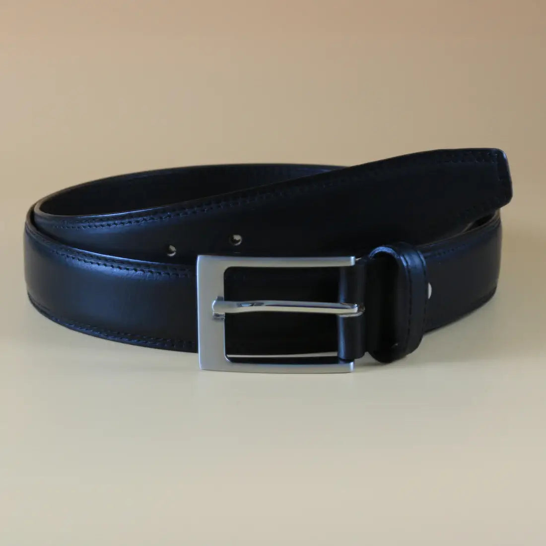 Black leather men’s belt with black stitching and silver rectangular buckle displayed on a clean light neutral background.