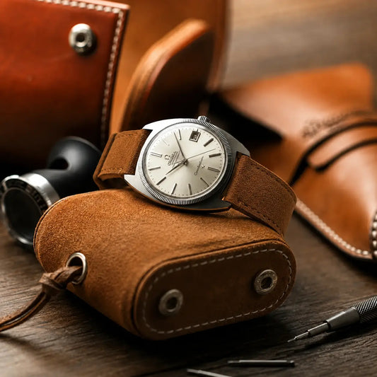 Omega Speedmaster chronograph paired with a premium brown suede leather strap, positioned on a black watch loupe against a warm blurred background for a refined luxury product scene.
