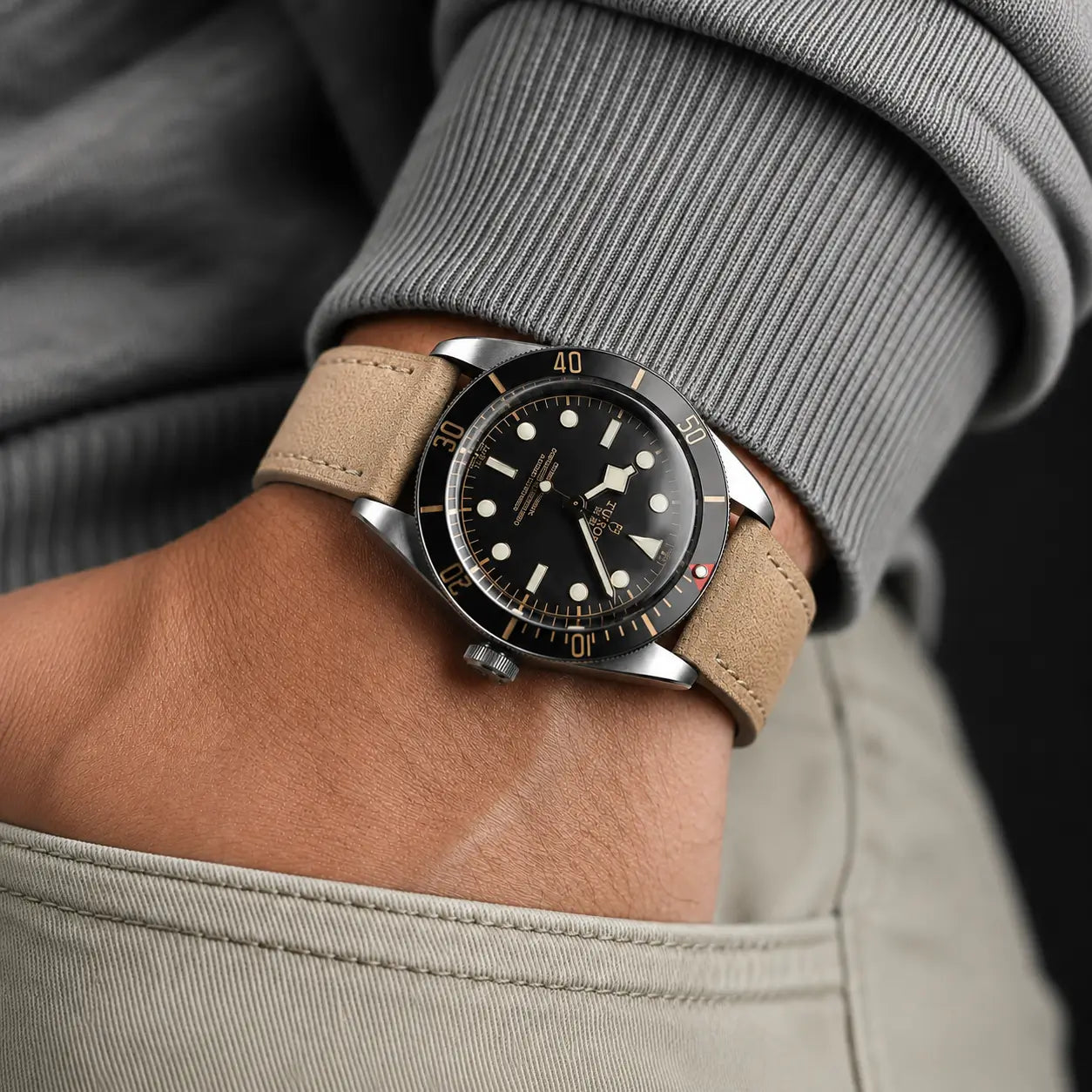 Lifestyle wrist shot featuring a vintage-inspired dive watch paired with a khaki suede leather strap. The watch is partially tucked into a trouser pocket with a grey ribbed sleeve, creating a casual premium look with soft natural lighting.