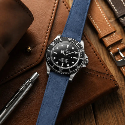 Premium lifestyle watch close-up showing a black Rolex-style diver watch fitted with a light blue suede leather strap. The watch rests diagonally across rich brown leather accessories with a metallic pen and warm ambient lighting, highlighting the soft suede texture and refined handcrafted aesthetic.